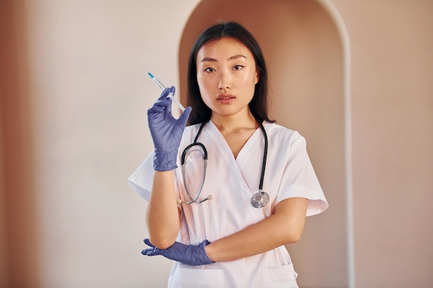 Médico con jeringa Joven mujer asiática seria de pie en el interior