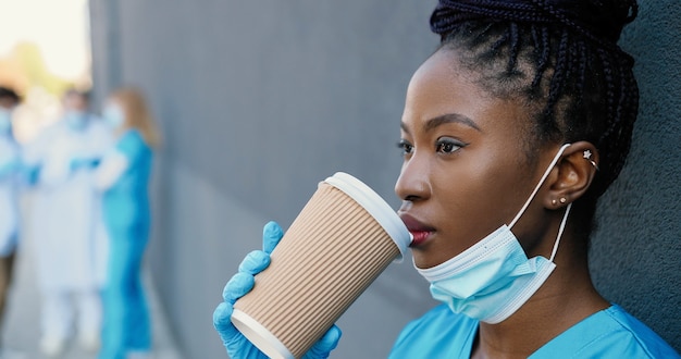 Médico jovem afro-americano cansado tirando a máscara médica e tomando uma bebida quente enquanto descansava e encostado na parede com os olhos fechados. linda enfermeira tomando café e descansando depois do trabalho duro