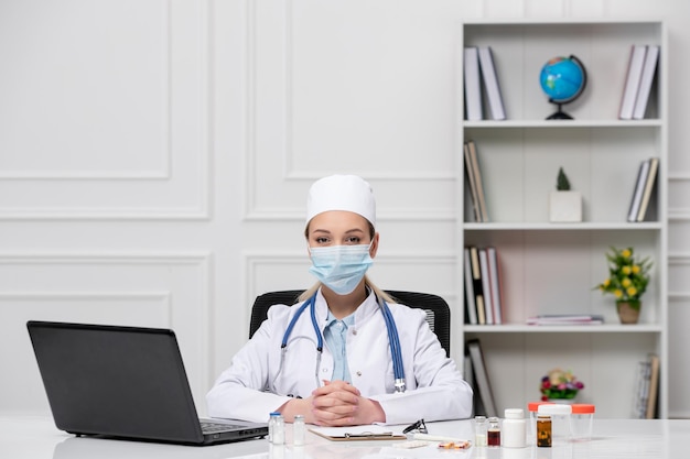 Médico jovem médico loiro de casaco branco e chapéu com computador usando máscara