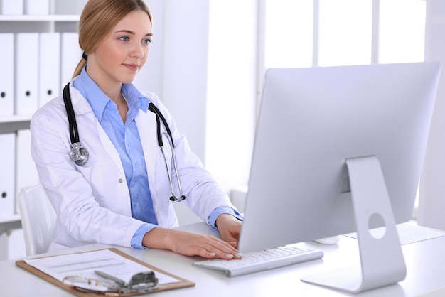 Médico jovem no trabalho no hospital olhando para monitor de pc desktop. Médico controla registros de histórico de medicação e resultados de exames. Conceito de medicina e saúde.