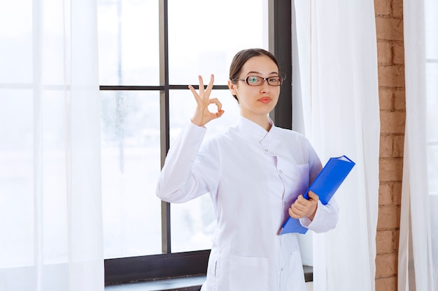 Médico joven en bata blanca con carpeta en sus manos cerca de la ventana.