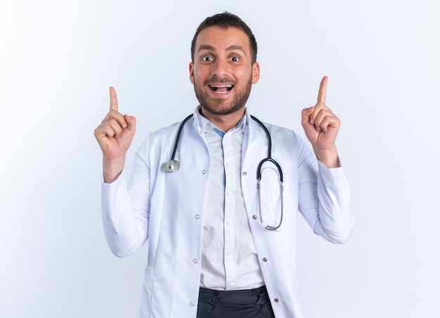 Médico joven en bata blanca y con estetoscopio alrededor del cuello feliz y positivo mostrando los dedos índices teniendo una gran idea de pie sobre la pared blanca