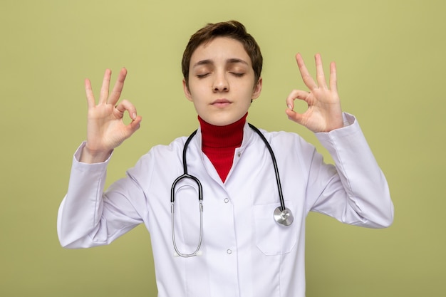 Médico joven en bata blanca con estetoscopio haciendo gesto de meditación tratando de relajarse con los ojos cerrados