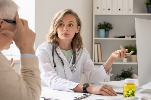 Médico joven confiado en bata blanca apuntando a la pantalla de la computadora mientras consulta a un paciente mayor y hace la presentación de un nuevo medicamento