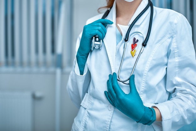 Médico joven hermosa en uniforme blanco y máscara en el rostro con un estetoscopio de cerca en el fondo de la clínica.