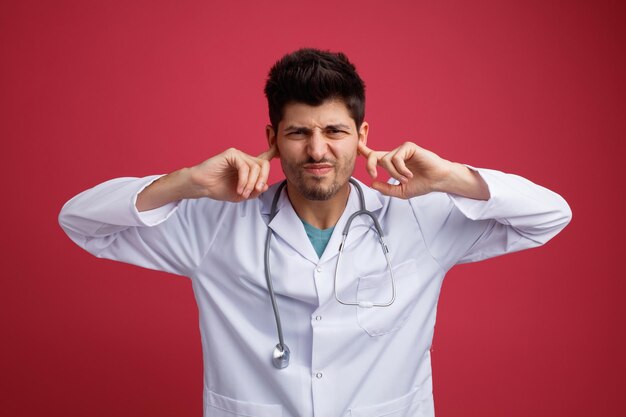 Médico joven irritado con uniforme médico y estetoscopio mirando a la cámara mientras mantiene los dedos en los oídos aislados en el fondo rojo