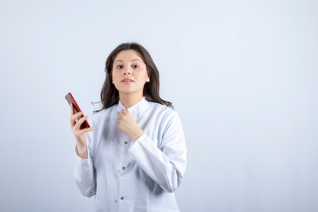 Médico joven que sostiene el teléfono celular en la pared blanca.