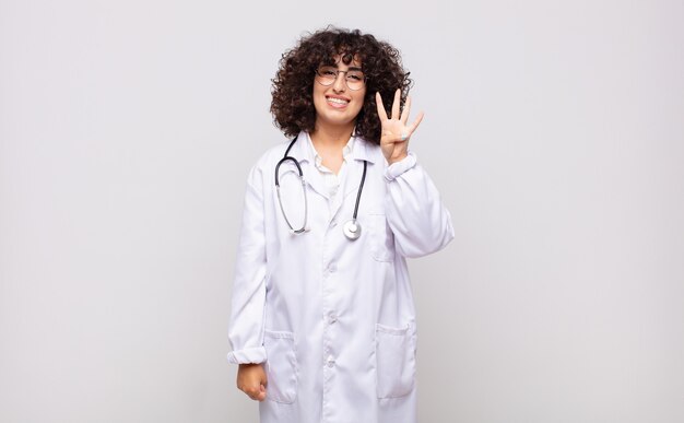 Médico joven sonriendo y mirando amigablemente, mostrando el número cuatro o cuarto con la mano hacia adelante, contando hacia atrás