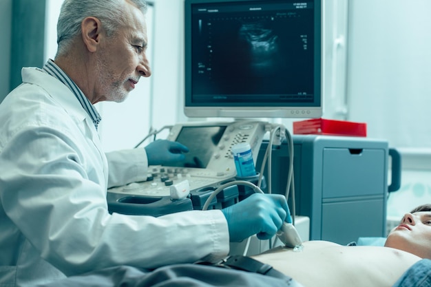 Médico maduro e amigável tocando a barriga de seu paciente com um transdutor de ultrassom e sorrindo levemente