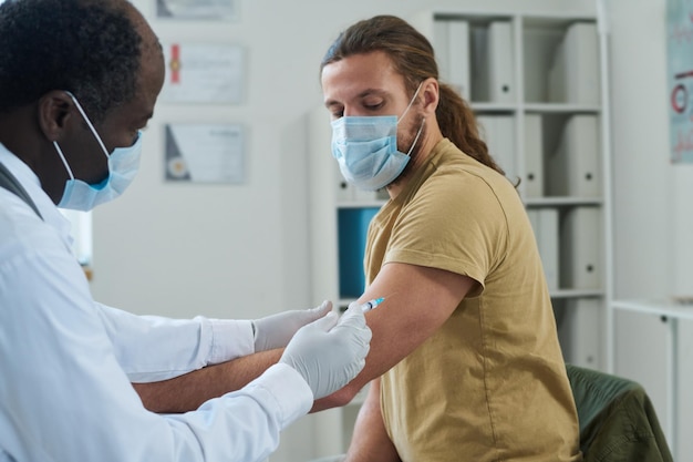 Médico maduro con máscara protectora que inyecta la vacuna a un paciente joven