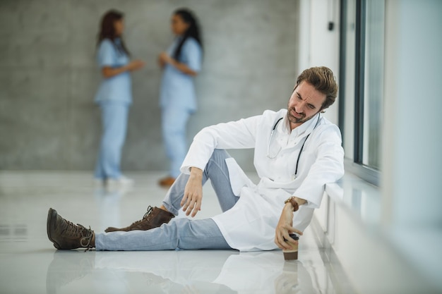 Un médico maduro preocupado sentado en el suelo cerca de una ventana mientras toma un café rápido en un pasillo del hospital durante la pandemia de Covid-19.