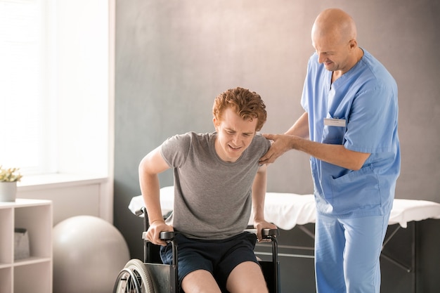 Médico maduro en uniforme ayudando a un joven con dolor a sentarse en una silla de ruedas después del entrenamiento de rehabilitación mientras sostiene su brazo y codo