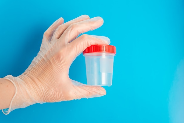 Médico mão na luva médica segura um recipiente de plástico vazio para testes em um fundo azul com espaço de cópia