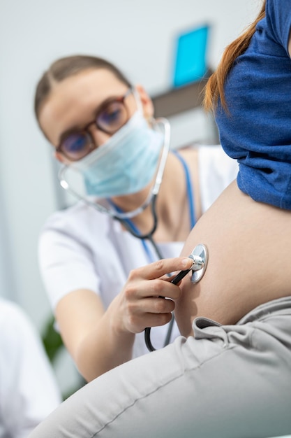 Un médico con una máscara examina el abdomen de una paciente embarazada con un estetoscopio