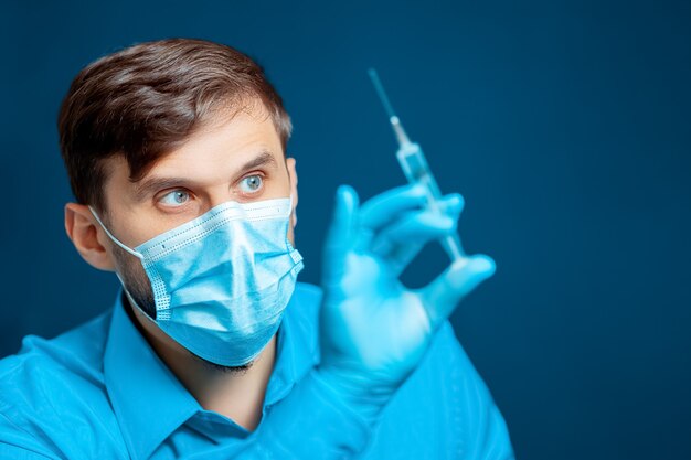 El médico con una máscara médica y guantes está vestido con un uniforme azul con una jeringa en la mano