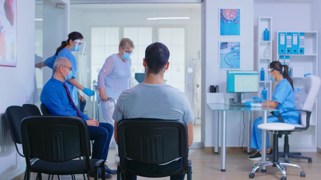 Médico con mascarilla contra el covid 19 invitando al paciente en la sala de examen. Enfermera ayudando a mujer mayor inválida con andador en la sala de espera del hospital. Sistema de atención médica durante una pandemia mundial