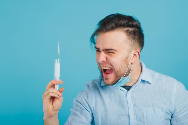 Médico masculino barbudo e segurando a seringa e gritando sobre fundo azul