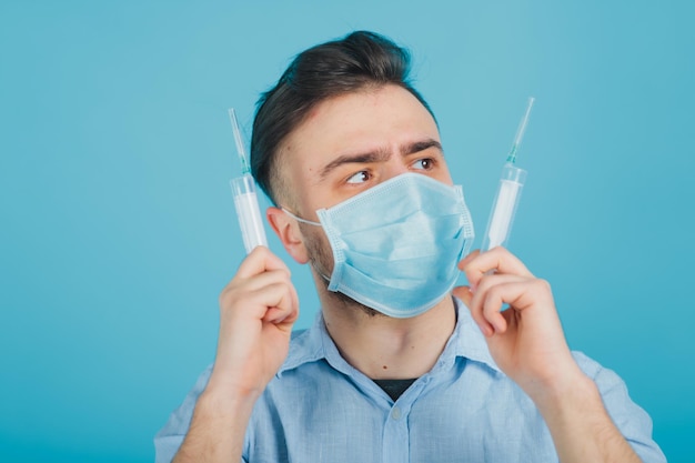 médico masculino barbudo em máscara segurando duas seringas nas mãos sobre fundo azul