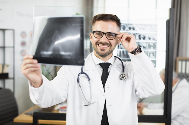 Médico masculino en bata de laboratorio y anteojos examinando escaneo de rayos x