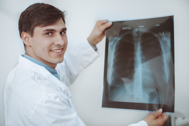 Médico masculino bonito alegre sorrindo para a câmera, examinando o raio-x do pulmão de um paciente