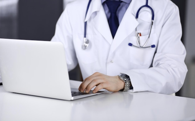 Médico masculino desconocido sentado y trabajando con una laptop en la clínica en su lugar de trabajo, de cerca. Médico joven en el trabajo. Servicio médico perfecto, concepto de medicina.