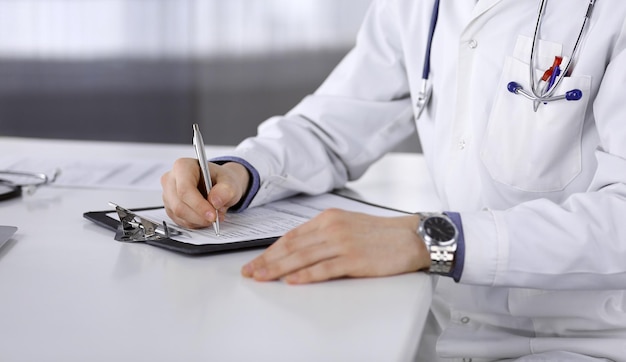 Médico masculino desconocido sentado y trabajando con el portapapeles del historial de medicamentos en la clínica en su lugar de trabajo, de cerca. Médico joven en el trabajo. Servicio médico perfecto, concepto de medicina.