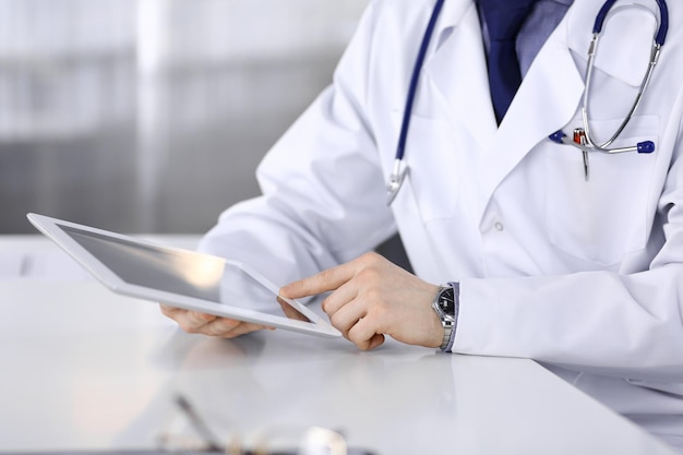 Médico masculino desconocido sentado y trabajando con una tableta en la clínica en su lugar de trabajo, primer plano. Médico joven en el trabajo. Servicio médico perfecto, concepto de medicina.