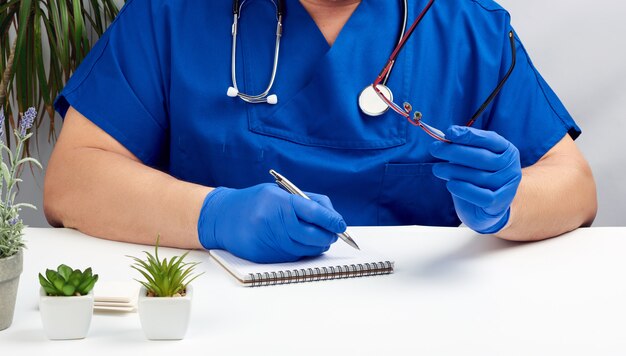 Médico masculino em um uniforme azul senta-se em uma mesa branca e escreve em um caderno de papel, usando luvas estéreis nas mãos