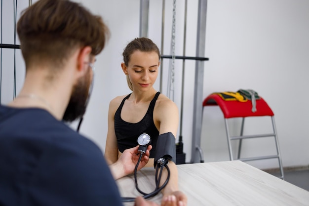 Médico masculino mide la presión de una joven deportista en el gimnasio para rehabilitación