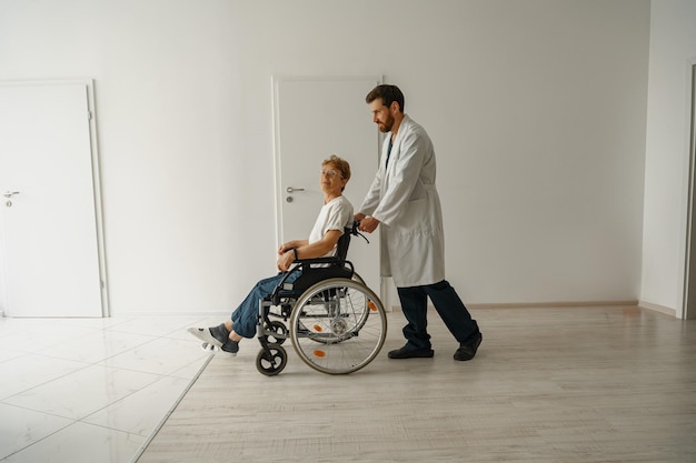 Médico masculino profesional que lleva a una paciente en silla de ruedas en el salón de la clínica de medicina