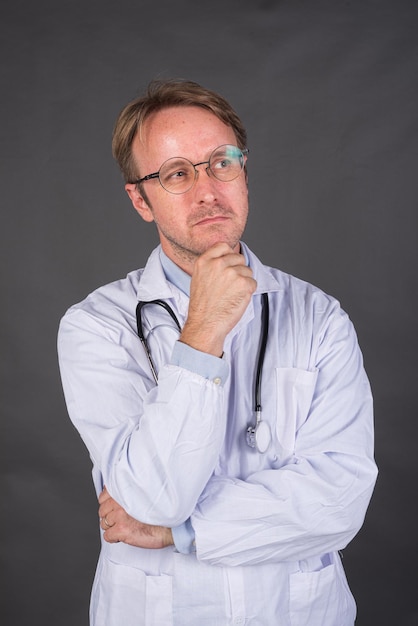 Médico masculino serio con estetoscopio sobre el cuello en abrigo médico pensando y mirando hacia arriba