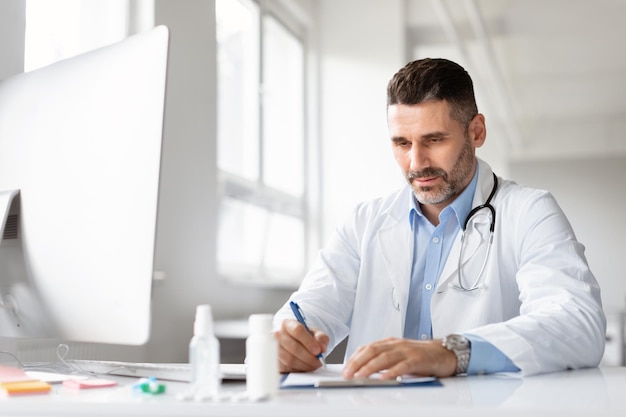 Foto médico masculino serio usando computadora y escribiendo notas en un diario médico sentado en el lugar de trabajo en la clínica