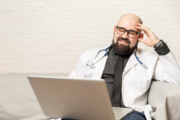 médico masculino se sienta en un sofá y trabaja frente a una computadora portátil. Trabajo remoto, consultas en línea durante la epidemia.