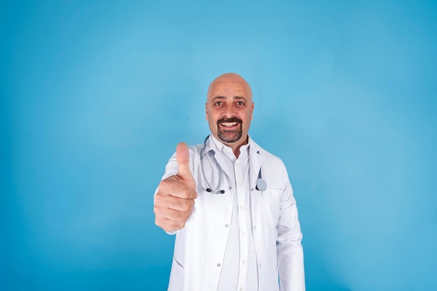 Médico masculino sonriente, con bata blanca y estetoscopio mostrando los pulgares hacia arriba con un fondo azul aislado.