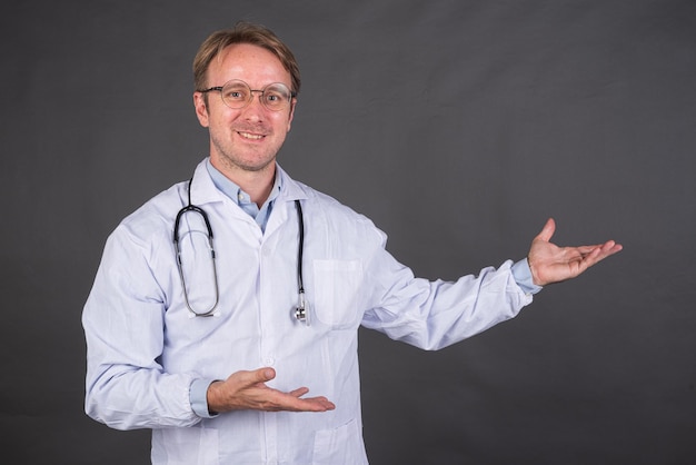 Médico masculino sorridente com estetoscópio em casaco médico gesticulando contra fundo cinza