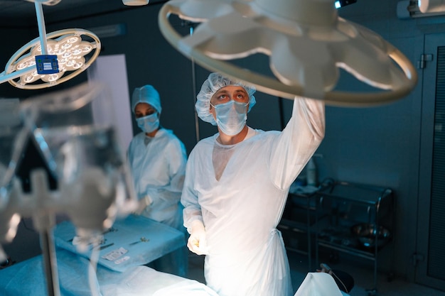 Médico masculino con uniformes quirúrgicos y máscaras ajustando la luz antes de la operación en la oscuridad