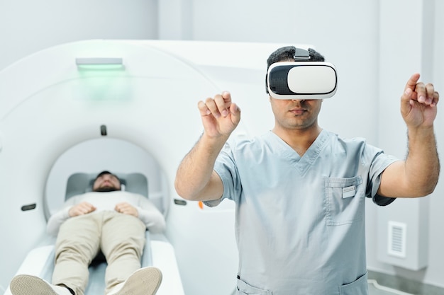 Médico mestiço contemporâneo em uniforme médico e fone de ouvido vr apontando para um grande display virtual contra o paciente e a máquina de ultra-som