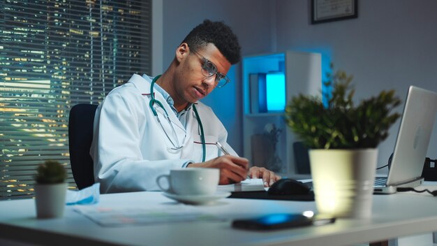 Médico mestiço fazendo anotações e conversando por videochamada no computador. Ele está trabalhando em casa por causa da pandemia de COVID-19.