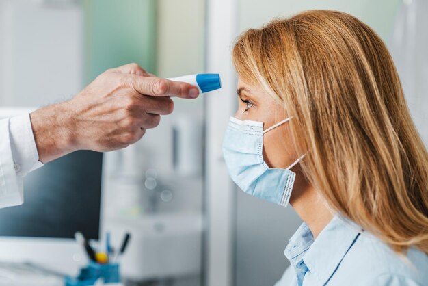 El médico mide la temperatura del paciente en la máscara con un termómetro sin contacto en la clínica