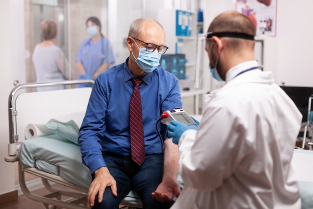 Médico midiendo la presión arterial del paciente mayor en la sala de consulta del hospital en el curso del examen covid19