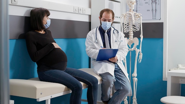 Médico y mujer embarazada consultando en el gabinete, discutiendo sobre atención médica infantil. Médico varón hablando con un paciente que espera un bebé, examen médico durante la pandemia de covid 19.