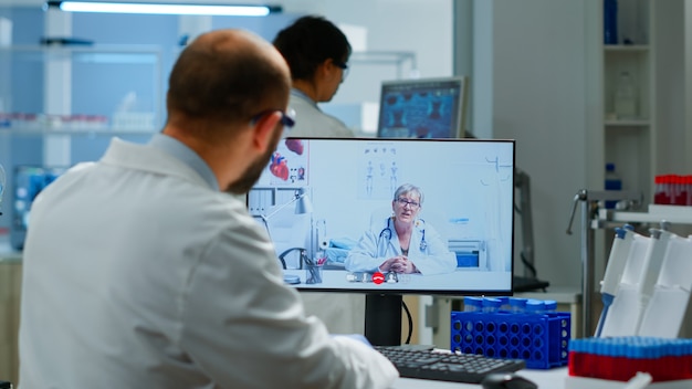 Médico de la mujer mayor que ofrece consejos médicos en línea al químico usando la cámara web de la PC. Científico sosteniendo una muestra de sangre durante una discusión en línea, conferencia virtual, ayudando en telemedicina, asistencia médica