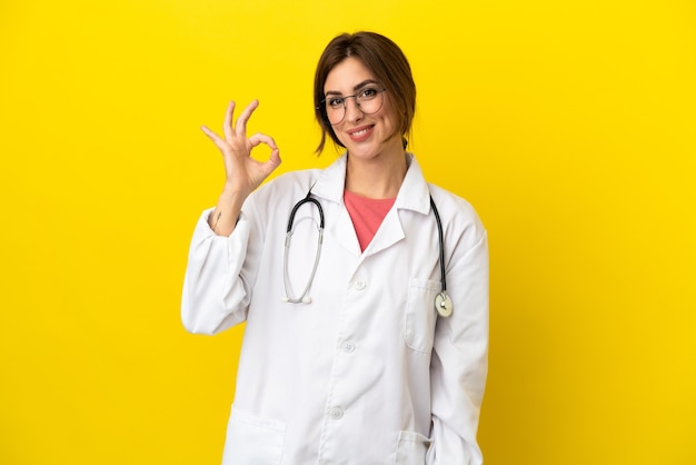 Médico mulher isolada em fundo amarelo mostrando sinal de ok com os dedos