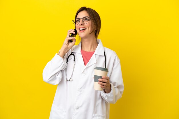 Médico mulher isolada em um fundo amarelo segurando um café para levar e um celular