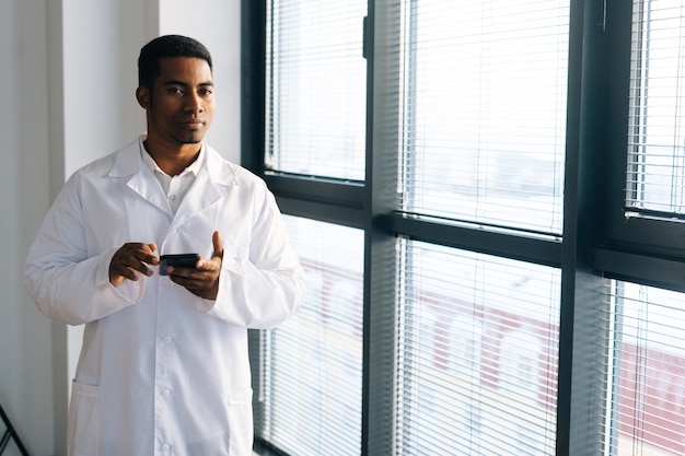 Médico negro sério vestindo casaco uniforme branco usando digitação no smartphone em pé pela janela no escritório do hospital