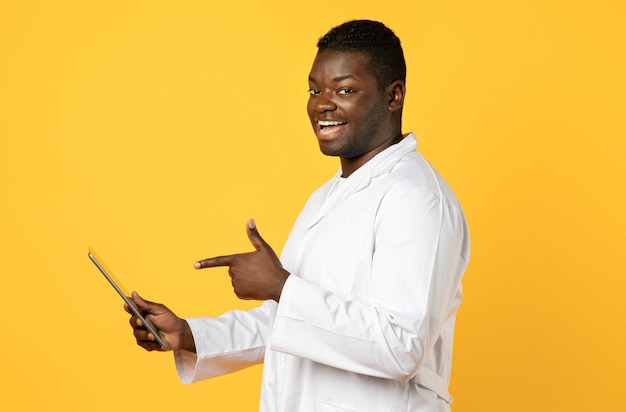 Un médico negro sonriente señalando una tableta digital sobre un fondo amarillo