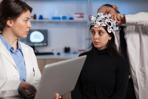 Médico neurologista segurando computador laptop mostrando tomografia para paciente mulher enquanto homem pesquisador ajustando fone de ouvido eeg durante experimento de neurociência. Equipe de pesquisadores analisando a evolução do cérebro