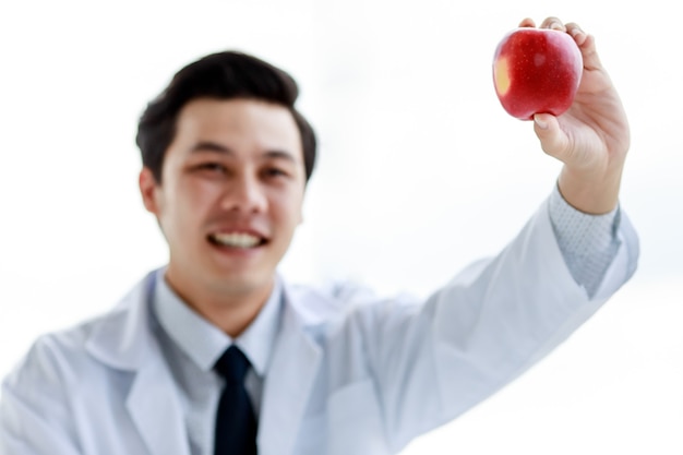 Médico nutricionista asiático joven atractivo con bata de laboratorio blanca y un estetoscopio sosteniendo una manzana roja sonriendo. Fondo blanco, aislado.