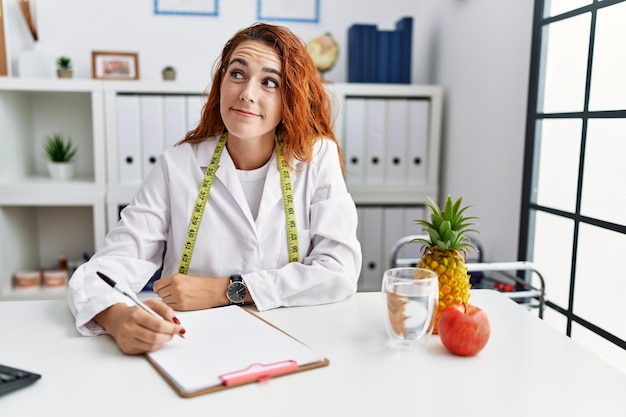 Médico nutricionista jovem ruiva na clínica sorrindo olhando para o lado e olhando para longe pensando.