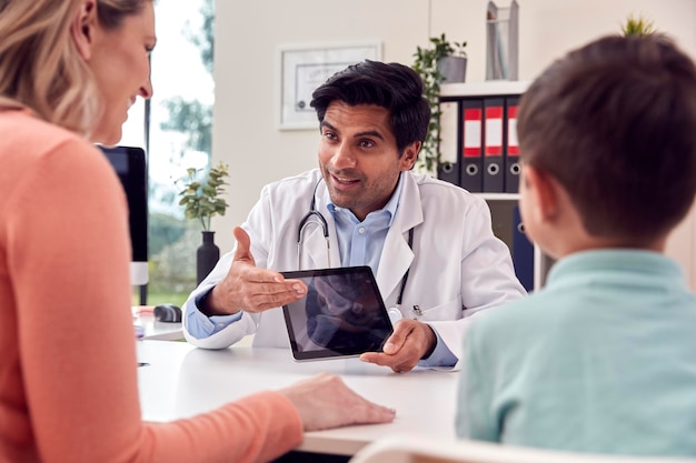 Médico o médico de cabecera en bata blanca que se encuentra con madre e hijo para una cita mirando el escaneo en la tableta digital